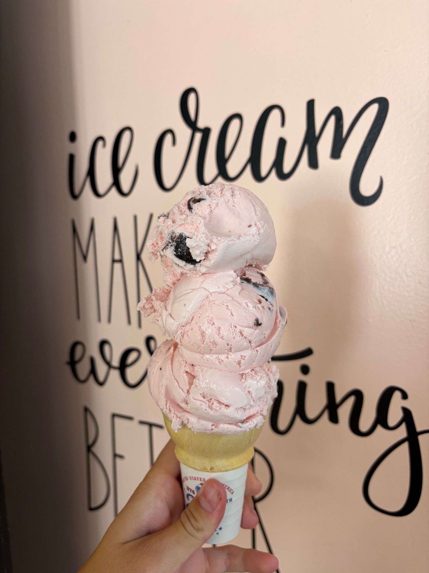 A three-scoop pink cone with chocolate chunks in front of the shop's "ice cream makes everything better" wall